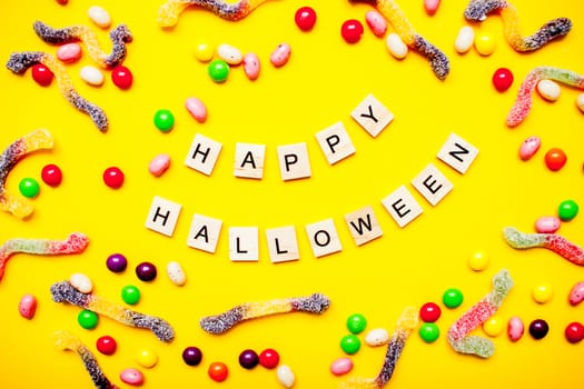 inscription from wooden blocks Happy Halloween and frame of multicolored candies and worms from gummy on a bright yellow background