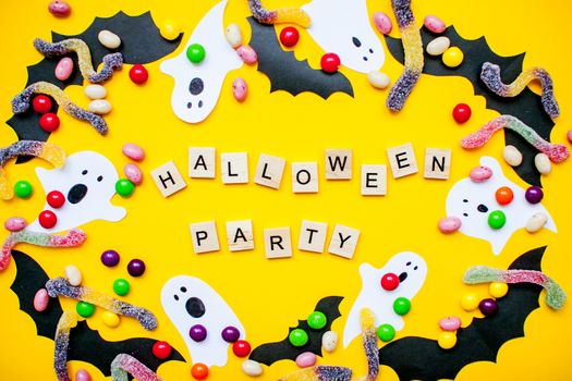 inscription from wooden blocks Halloween party and frame made of paper homemade bats and paper ghosts and multicolored candies and worms from gummy on a bright yellow background