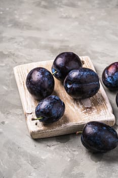 Fresh ripe plum fruits with water drops on wooden cutting board, stone concrete background, angle view