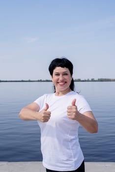 Sport and fitness. Senior sport. Active seniors. Active and happy senior woman in sportswear showing thumbs up working out near the riverside