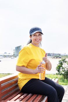 Sport and fitness. Senior sport. Active seniors. Smiling senior woman drinking water after workout outdoors on the sports ground showing thumbs up