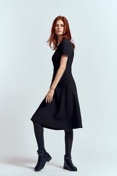 A woman in a black dress on a light background and pantyhose shoes red hair and pose in full growth. High quality photo