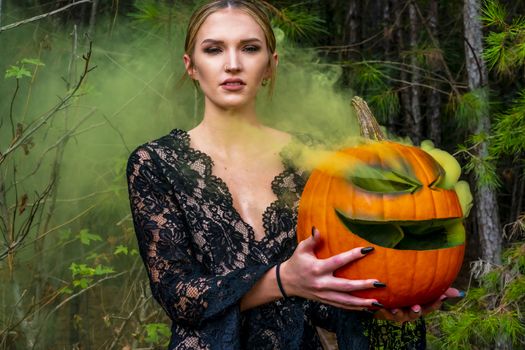 A beautiful blonde model wearing all black poses with a smoking pumpkin in an outdoor environment