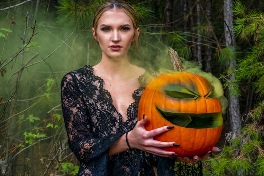 A beautiful blonde model wearing all black poses with a smoking pumpkin in an outdoor environment