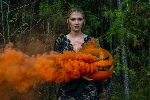 A beautiful blonde model wearing all black poses with a smoking pumpkin in an outdoor environment