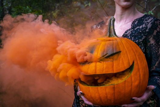 A beautiful blonde model wearing all black poses with a smoking pumpkin in an outdoor environment