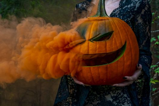 A beautiful blonde model wearing all black poses with a smoking pumpkin in an outdoor environment