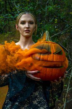 A beautiful blonde model wearing all black poses with a smoking pumpkin in an outdoor environment