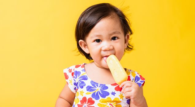 Happy portrait Asian baby or kid cute little girl attractive laugh smile wearing dick pattern shirt holds and eating sweet wooden ice cream, studio shot isolated on yellow background, summer concept