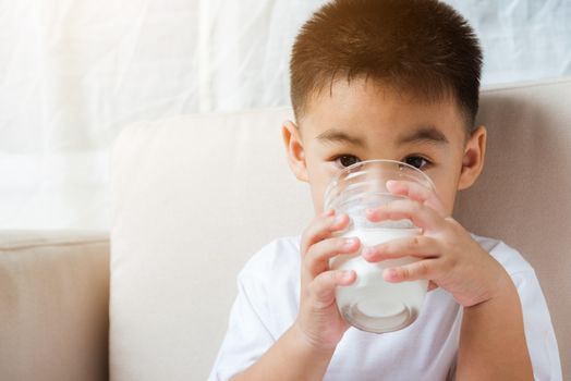 Close up of happy Asian little cute child boy hand holding milk glass he drinking white milk during sitting on the sofa at home after lunch. Daily life health care Medicine food