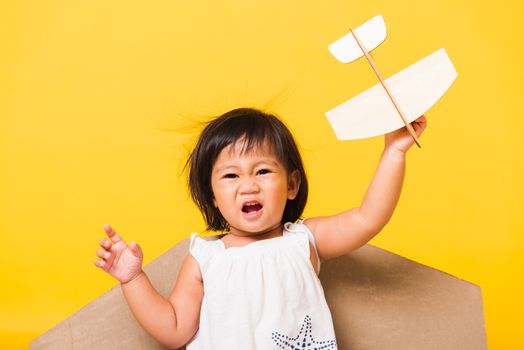 Happy Asian beautiful funny baby little girl smile wear pilot hat play with toy cardboard airplane wings fly hold plane toy, studio shot isolated yellow background, Startup freedom concept
