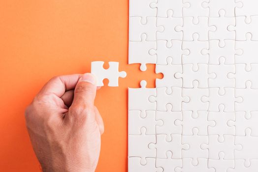 Top view flat lay of hand-holding last piece white paper jigsaw puzzle game last pieces put to place for solve problem complete mission, studio shot on an orange background, quiz calculation concept