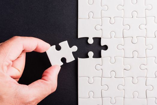 Top view flat lay of hand-holding last piece white paper jigsaw puzzle game last pieces put to place for solve problem complete mission, studio shot on a black background, quiz calculation concept