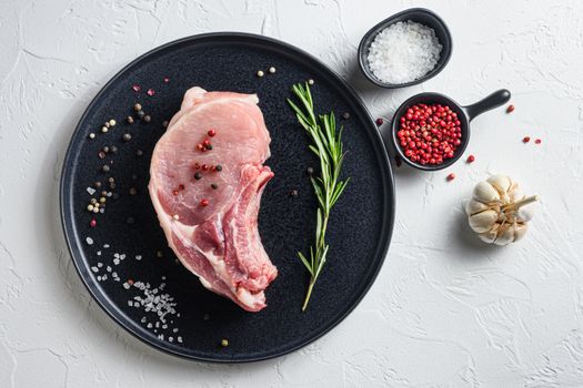 Raw Pork Loin chops in a black round plate on a white stone background with rosemary garlic peppercorns ingredients for grill top view.