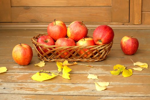 Autumn still life with ripe red apples in a wicker basket and yellow leaves scattered on a wooden surface painted brown