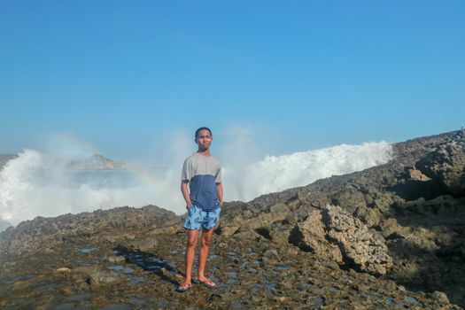 Teenager in shorts and a T-shirt is standing on a cliff.. Waves crashing over a rocky on ocean wave background. Young asian man standing on the rock.