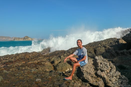 A man sits on a stone by the ocean. Waves breaking on a rock cliff. Teenager by the stormy sea. Young guy on a cliff with a sea view. A young Asian man in shorts and a T-shirt sitting on a cliff.