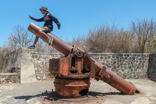 A young guy sitting on historic, ancient cannon. Mirjam Jepan on Lombok Island, Indonesia. Asian teenager sitting on an old rusty cannon in a stone fortification.