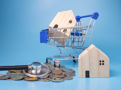 A model wooden house on a shopping cart With a pile of coins and stethoscope On a blue background. Mortgage concept. Money and house. Financial status.

