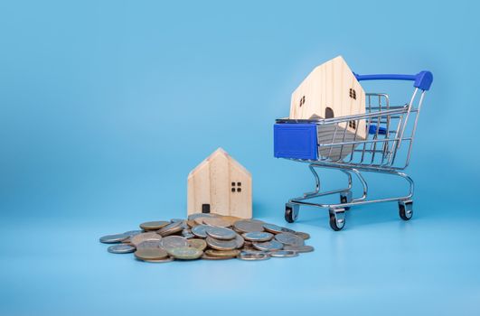 A model wooden house on a shopping cart With a pile of coins On a blue background. Mortgage concept. Money and house.