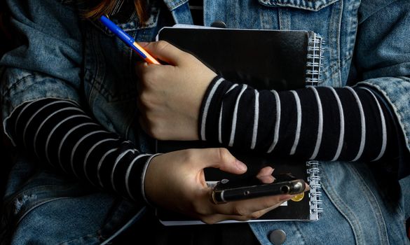 Girl's hands.She clutches black notebooks and an orange pen in one hand, and flips through her phone with the other. The concept of the business school