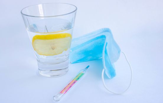 A glass of water with lemon, a mask and a thermometer on a white background. Glass mercury thermometer with high temperature