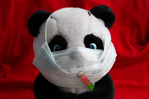 A toy panda in a medical mask with a thermometer sits on a red background. Coronavirus treatment concept