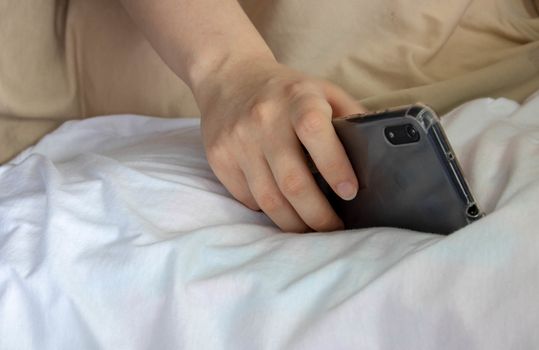 Woman hand under blanket being woken by mobile phone in bedroom