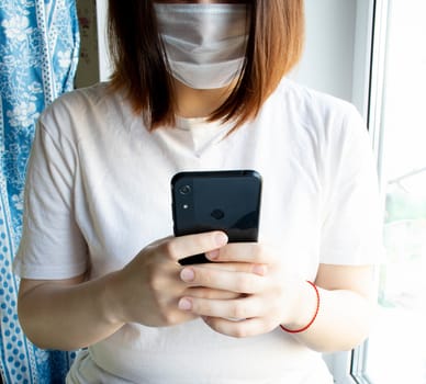 Medical mask on the face of an unknown girl, hands and mobile phone.The house near the