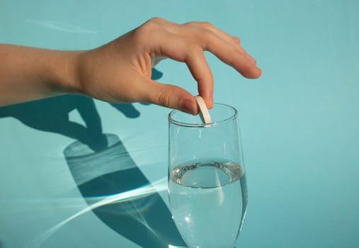 Against a blue background, a hand drops a dissolving fizzy aspirin tablet into a glass of water