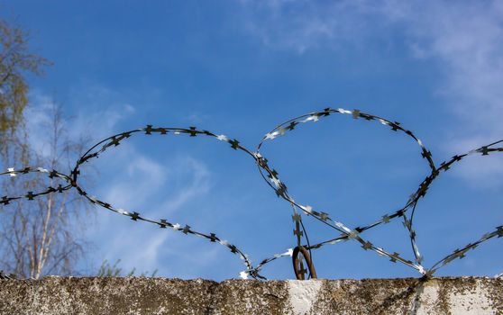 A heart made of barbed wire against a blue sky on a concrete fence.The object is protected.