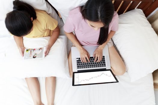 Top view Asian mother and daugther looking laptop and tablet on bed in bedroom. Business women woking with laptop at home. Girl enjoy speak with father video call  on bed. soft focus photo