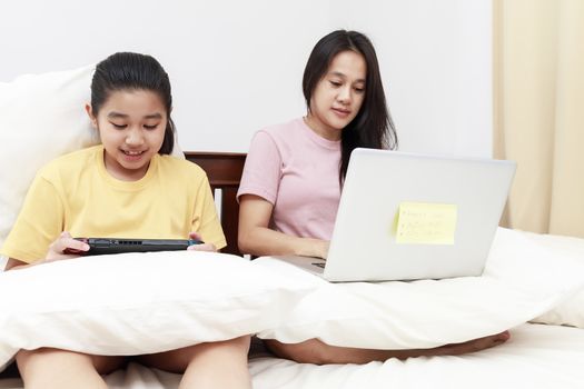 Asian mother and daugther looking laptop and game console on bed in bedroom. Business women woking with laptop at home with her daugther smiling enjoy with game console on bed. Work at home concept.