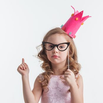 Funny princess girl in pink dress and crown holding party glasses on stick and saying something smart with finger up