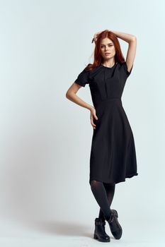 A woman in a black dress on a light background and pantyhose shoes red hair and pose in full growth. High quality photo