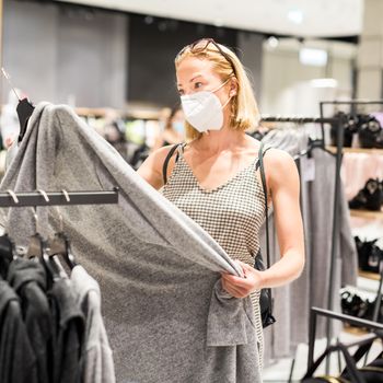 Fashionable woman wearing protective face mask shopping clothes in reopen retail shopping store. New normal lifestyle during corona virus pandemic.