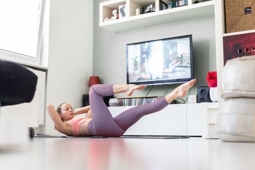 Attractive sporty woman working out at home, doing pilates exercise in front of television in small studio appartment. Social distancing. Stay healthy and stay at home during corona virus pandemic.