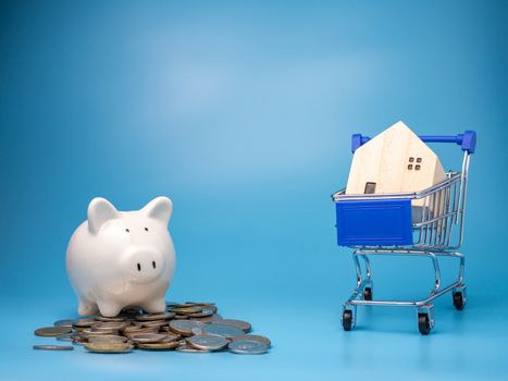 A model wooden house on a shopping cart With a pile of coins and Piggy bank  On a blue background. Mortgage concept. Money and house. Financial status.