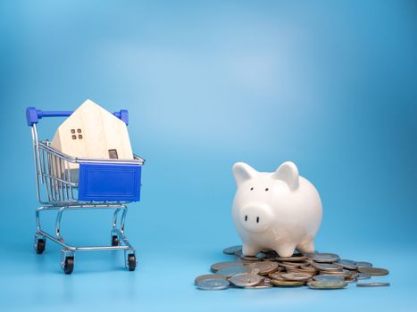 A model wooden house on a shopping cart With a pile of coins and Piggy bank  On a blue background. Mortgage concept. Money and house. Financial status.