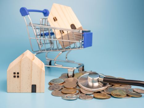 A model wooden house on a shopping cart With a pile of coins and stethoscope On a blue background. Mortgage concept. Money and house. Financial status.