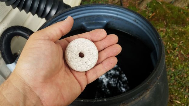 hand holding circular white mosquito tablet insecticide over rain barrel with water