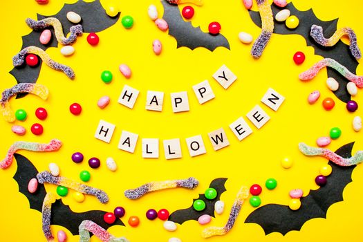 inscription from wooden blocks Happy Halloween and frame made of paper homemade bats and multicolored candies and worms from gummy on a bright yellow background
