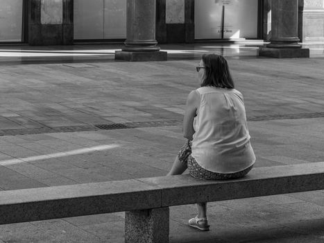 Woman sitting on a bench shot from behind in a street in the center of Milan, street photography in black and white