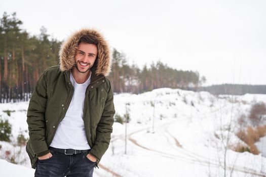Handsome bearded young caucasian man standing outdoors fur hood in winter season forest. Attractive stylish european guy walking snowy christmas woodland Season holiday leisure