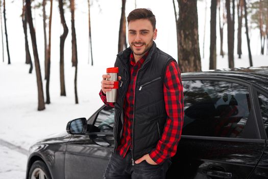 Attractive Caucasian man stands winter forest near car drink hot beverage thermos. Winter holiday road trip. Cold season. Portrait young handsome unshaved driver dressed red checkered shirt vest
