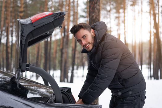 Young adult handsome man standing winter forest near his car. Attractive caucasian guy waiting for help when car is broken. Winter landscape road trip.