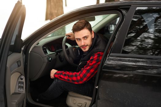 Young adult attractive Caucasian man sits at the wheel of his car sunny winter day. Wintertime road trip. Happy smiling hipster guy sitting in car and looking window. Portrait Positive driver