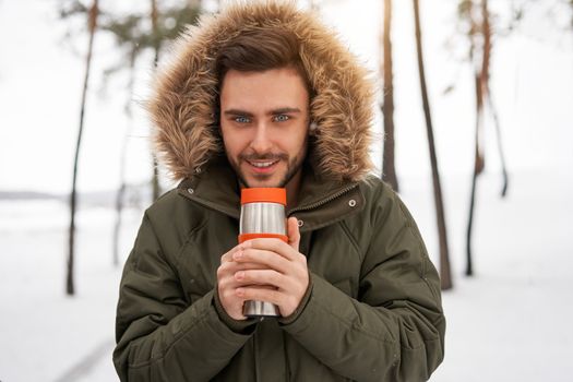 One Attractive Caucasian man stands winter forest drink hot beverage thermos. Winter holiday leisure. Cold season. Portrait young handsome unshaved Modern hipster guy traveling walking in winter park