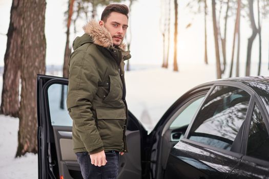 Attractive Caucasian man stands in winter forest near his car. Winter holiday road trip. Cold season. Portrait young handsome unshaved driver. Modern hipster guy traveling on car.