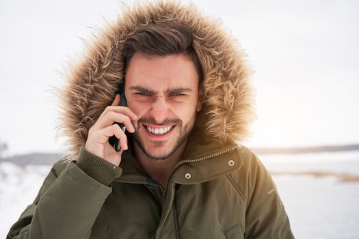 Handsome smiling brutal bearded young caucasian man standing outdoors fur hood in winter season forest Talking on a cell phone Attractive stylish european guy walking snowy christmas landscape